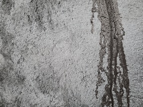 Rough textured concrete surface with darker patches and vertical streaks indicating water or moisture exposure. The uneven texture creates a rugged and industrial appearance.