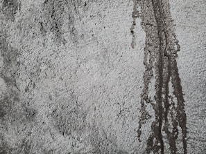 Rough textured concrete surface with darker patches and vertical streaks indicating water or moisture exposure. The uneven texture creates a rugged and industrial appearance.