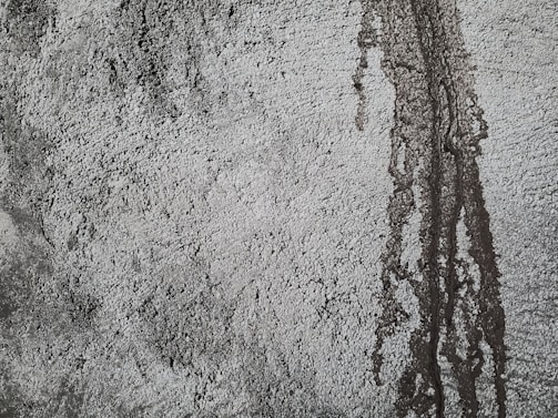 Rough textured concrete surface with darker patches and vertical streaks indicating water or moisture exposure. The uneven texture creates a rugged and industrial appearance.