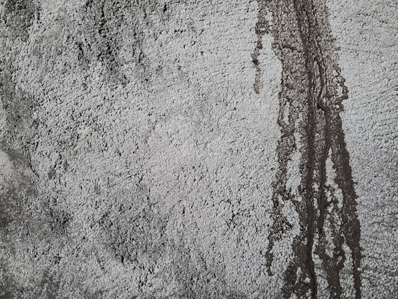 Rough textured concrete surface with darker patches and vertical streaks indicating water or moisture exposure. The uneven texture creates a rugged and industrial appearance.