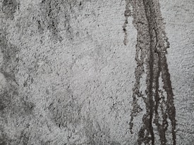 Rough textured concrete surface with darker patches and vertical streaks indicating water or moisture exposure. The uneven texture creates a rugged and industrial appearance.