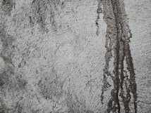 Rough textured concrete surface with darker patches and vertical streaks indicating water or moisture exposure. The uneven texture creates a rugged and industrial appearance.