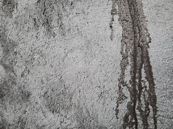 Rough textured concrete surface with darker patches and vertical streaks indicating water or moisture exposure. The uneven texture creates a rugged and industrial appearance.