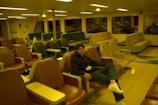 A person is sitting in a nearly empty room filled with various styles of seating. The seats range in color from brown to green and appear to be arranged in an orderly fashion. The lighting has a warm yellow tint, and there are large windows along the side, reflecting some of the chairs.