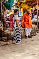 An outdoor market scene where an elderly man wearing a bright orange traditional outfit and holding flowers is engaged in conversation with a woman in a striped saree. They are standing near a stone platform with scattered sandals and surrounded by colorful fabric canopies.