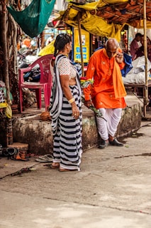 An outdoor market scene where an elderly man wearing a bright orange traditional outfit and holding flowers is engaged in conversation with a woman in a striped saree. They are standing near a stone platform with scattered sandals and surrounded by colorful fabric canopies.