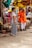 An outdoor market scene where an elderly man wearing a bright orange traditional outfit and holding flowers is engaged in conversation with a woman in a striped saree. They are standing near a stone platform with scattered sandals and surrounded by colorful fabric canopies.