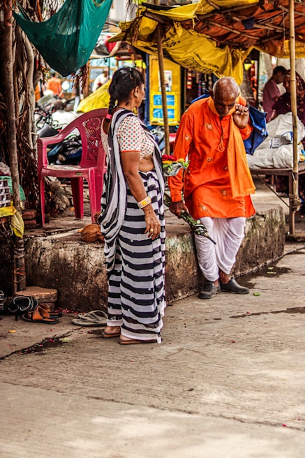 An outdoor market scene where an elderly man wearing a bright orange traditional outfit and holding flowers is engaged in conversation with a woman in a striped saree. They are standing near a stone platform with scattered sandals and surrounded by colorful fabric canopies.