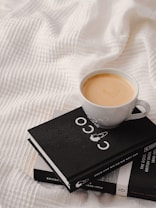 A neatly placed cup of coffee sits atop two stacked books on a textured white fabric, creating a cozy and inviting scene. The cup is filled with creamy coffee, and the visible book has a stylish black cover featuring a minimalist design.