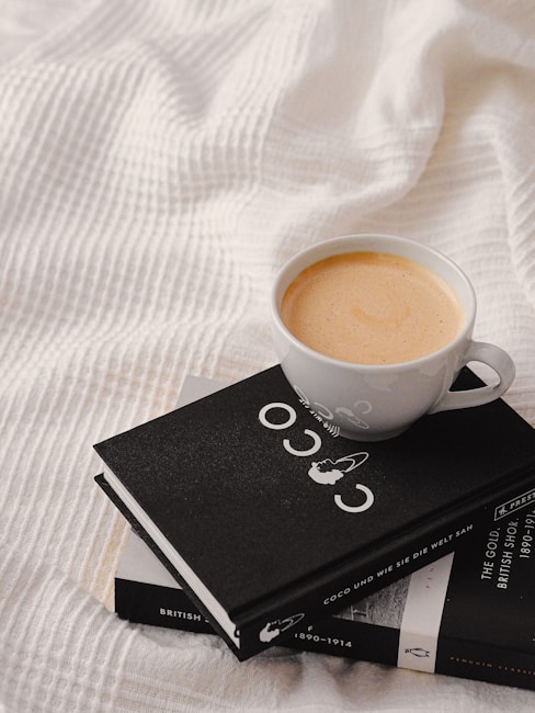 A neatly placed cup of coffee sits atop two stacked books on a textured white fabric, creating a cozy and inviting scene. The cup is filled with creamy coffee, and the visible book has a stylish black cover featuring a minimalist design.