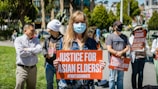 People are gathered outdoors holding signs with messages advocating for justice for Asian elders and against Asian hate. The central figure is a person wearing a face mask and holding a sign that reads 'Justice for Asian Elders'. The setting appears to be a public park with trees and buildings in the background.