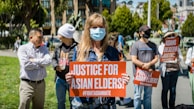People are gathered outdoors holding signs with messages advocating for justice for Asian elders and against Asian hate. The central figure is a person wearing a face mask and holding a sign that reads 'Justice for Asian Elders'. The setting appears to be a public park with trees and buildings in the background.