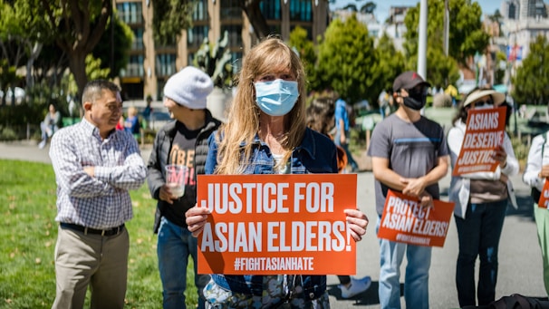 People are gathered outdoors holding signs with messages advocating for justice for Asian elders and against Asian hate. The central figure is a person wearing a face mask and holding a sign that reads 'Justice for Asian Elders'. The setting appears to be a public park with trees and buildings in the background.
