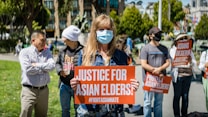 People are gathered outdoors holding signs with messages advocating for justice for Asian elders and against Asian hate. The central figure is a person wearing a face mask and holding a sign that reads 'Justice for Asian Elders'. The setting appears to be a public park with trees and buildings in the background.