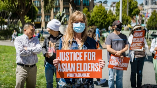 People are gathered outdoors holding signs with messages advocating for justice for Asian elders and against Asian hate. The central figure is a person wearing a face mask and holding a sign that reads 'Justice for Asian Elders'. The setting appears to be a public park with trees and buildings in the background.