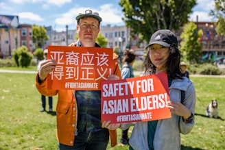 Two individuals holding signs with messages addressing anti-Asian hate, standing in a park. The setting is sunny, and there are trees and buildings in the background. The signs have bold text in both English and another language, emphasizing safety and justice.