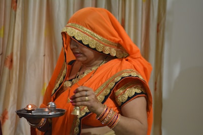 A woman dressed in traditional orange attire, including a saree and blouse with gold embroidery, holds a small metal platter with lit candles and a bell. She appears engaged in a religious ritual or ceremony. The background features a curtain with subtle floral patterns.