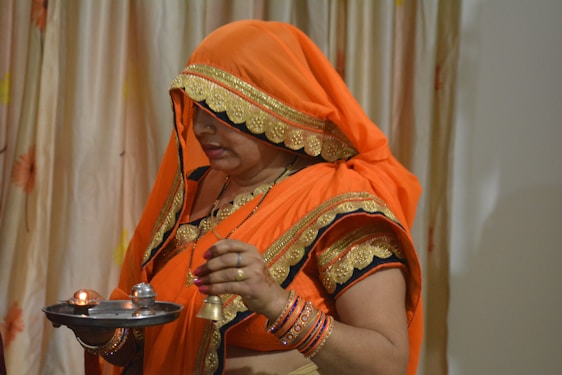 A woman dressed in traditional orange attire, including a saree and blouse with gold embroidery, holds a small metal platter with lit candles and a bell. She appears engaged in a religious ritual or ceremony. The background features a curtain with subtle floral patterns.