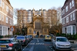 A narrow street lined with parked cars leads to a large, gothic-style church with a prominent arched window and spire. Tall apartment buildings loom in the background, and bare trees flank the street. A school sign is visible, and various vehicles, including a police car, are parked along the curb.