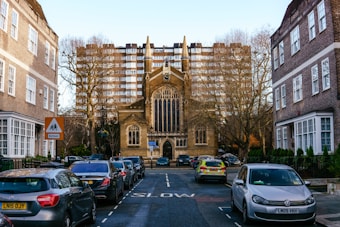A narrow street lined with parked cars leads to a large, gothic-style church with a prominent arched window and spire. Tall apartment buildings loom in the background, and bare trees flank the street. A school sign is visible, and various vehicles, including a police car, are parked along the curb.