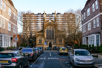 A narrow street lined with parked cars leads to a large, gothic-style church with a prominent arched window and spire. Tall apartment buildings loom in the background, and bare trees flank the street. A school sign is visible, and various vehicles, including a police car, are parked along the curb.
