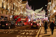 a group of people walk down a sidewalk next to a street with cars and christmas lights