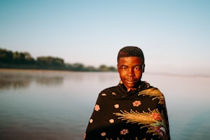 A person stands in front of a calm body of water with a shawl wrapped around their shoulders. The shawl features floral and intricate patterns. The background includes a serene landscape with distant trees under a clear blue sky.