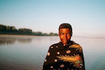 A person stands in front of a calm body of water with a shawl wrapped around their shoulders. The shawl features floral and intricate patterns. The background includes a serene landscape with distant trees under a clear blue sky.