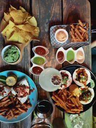 A variety of dishes are arranged on a wooden table. There is a wooden bowl filled with crispy tortilla chips alongside a bowl of guacamole. Multiple small dishes contain different colored sauces, including red and green ones. Two large plates contain tacos topped with fresh salsa, sour cream, and herbs, accompanied by sweet potato fries and a corn cob. A cocktail with a dried citrus garnish and a glass of wine are also visible.