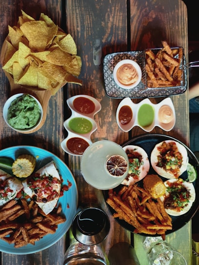 A variety of dishes are arranged on a wooden table. There is a wooden bowl filled with crispy tortilla chips alongside a bowl of guacamole. Multiple small dishes contain different colored sauces, including red and green ones. Two large plates contain tacos topped with fresh salsa, sour cream, and herbs, accompanied by sweet potato fries and a corn cob. A cocktail with a dried citrus garnish and a glass of wine are also visible.