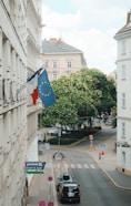 The image depicts a city street scene with a European building adorned with flags, including the French and European Union flags. The street below is calm, featuring parked cars, a bicycle, and various street signs. In the background, there is a lush, green tree and more buildings under a partly cloudy sky.