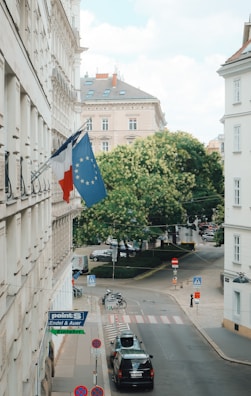 The image depicts a city street scene with a European building adorned with flags, including the French and European Union flags. The street below is calm, featuring parked cars, a bicycle, and various street signs. In the background, there is a lush, green tree and more buildings under a partly cloudy sky.