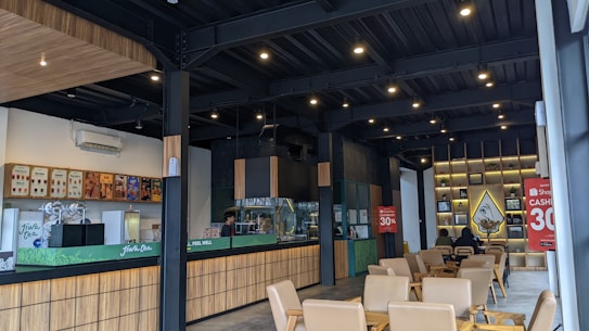 A modern, warmly lit caf&eacute; with wooden accents, featuring a counter displaying various coffee options. The seating area includes several beige chairs and tables. The walls are decorated with shelves holding small plants and art pieces. Two large promotional signs indicating a 30% discount are visible.