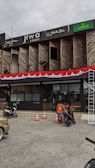 A commercial building with multiple businesses, including Jiwa Toast and Jiwa Tea, displayed on the upper facade. The exterior is designed with wooden panels and decorative red and white bunting. Two men are working on a window ledge, and below, a person with a safety vest is near two motorcycles. The foreground shows traffic cones and part of a parked car.