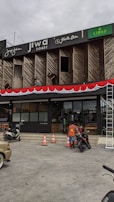 A commercial building with multiple businesses, including Jiwa Toast and Jiwa Tea, displayed on the upper facade. The exterior is designed with wooden panels and decorative red and white bunting. Two men are working on a window ledge, and below, a person with a safety vest is near two motorcycles. The foreground shows traffic cones and part of a parked car.