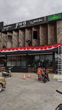 A commercial building with multiple businesses, including Jiwa Toast and Jiwa Tea, displayed on the upper facade. The exterior is designed with wooden panels and decorative red and white bunting. Two men are working on a window ledge, and below, a person with a safety vest is near two motorcycles. The foreground shows traffic cones and part of a parked car.
