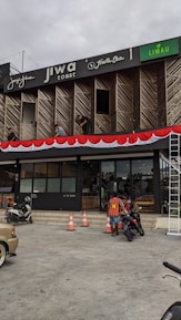 A commercial building with multiple businesses, including Jiwa Toast and Jiwa Tea, displayed on the upper facade. The exterior is designed with wooden panels and decorative red and white bunting. Two men are working on a window ledge, and below, a person with a safety vest is near two motorcycles. The foreground shows traffic cones and part of a parked car.
