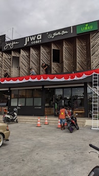 A commercial building with multiple businesses, including Jiwa Toast and Jiwa Tea, displayed on the upper facade. The exterior is designed with wooden panels and decorative red and white bunting. Two men are working on a window ledge, and below, a person with a safety vest is near two motorcycles. The foreground shows traffic cones and part of a parked car.