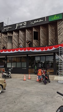 A commercial building with multiple businesses, including Jiwa Toast and Jiwa Tea, displayed on the upper facade. The exterior is designed with wooden panels and decorative red and white bunting. Two men are working on a window ledge, and below, a person with a safety vest is near two motorcycles. The foreground shows traffic cones and part of a parked car.