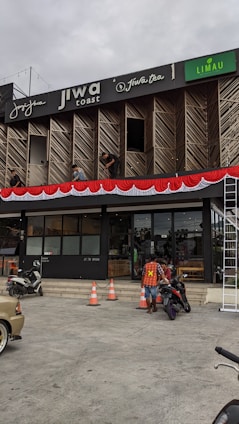 A commercial building with multiple businesses, including Jiwa Toast and Jiwa Tea, displayed on the upper facade. The exterior is designed with wooden panels and decorative red and white bunting. Two men are working on a window ledge, and below, a person with a safety vest is near two motorcycles. The foreground shows traffic cones and part of a parked car.