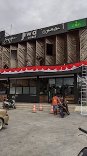 A commercial building with multiple businesses, including Jiwa Toast and Jiwa Tea, displayed on the upper facade. The exterior is designed with wooden panels and decorative red and white bunting. Two men are working on a window ledge, and below, a person with a safety vest is near two motorcycles. The foreground shows traffic cones and part of a parked car.