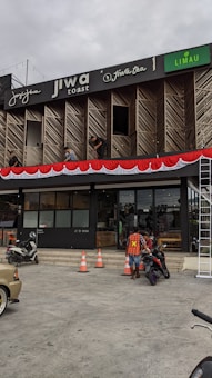 A commercial building with multiple businesses, including Jiwa Toast and Jiwa Tea, displayed on the upper facade. The exterior is designed with wooden panels and decorative red and white bunting. Two men are working on a window ledge, and below, a person with a safety vest is near two motorcycles. The foreground shows traffic cones and part of a parked car.