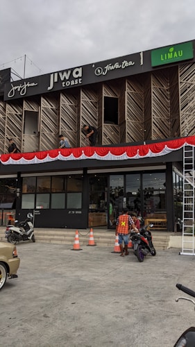 A commercial building with multiple businesses, including Jiwa Toast and Jiwa Tea, displayed on the upper facade. The exterior is designed with wooden panels and decorative red and white bunting. Two men are working on a window ledge, and below, a person with a safety vest is near two motorcycles. The foreground shows traffic cones and part of a parked car.