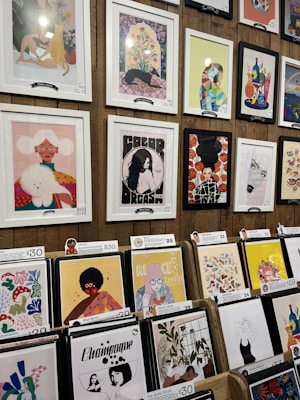A display of various framed and unframed art pieces on a wooden wall and stands. The artworks include colorful illustrations, abstract designs, and portraits featuring animals, people, and plants. Some pieces are priced at $68 and $69, while others are priced around $29 to $30.