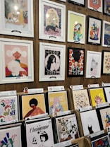 A display of various framed and unframed art pieces on a wooden wall and stands. The artworks include colorful illustrations, abstract designs, and portraits featuring animals, people, and plants. Some pieces are priced at $68 and $69, while others are priced around $29 to $30.