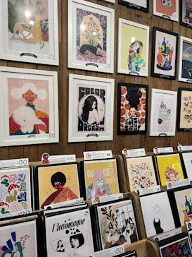 A display of various framed and unframed art pieces on a wooden wall and stands. The artworks include colorful illustrations, abstract designs, and portraits featuring animals, people, and plants. Some pieces are priced at $68 and $69, while others are priced around $29 to $30.