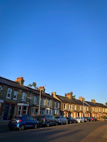 A row of traditional terraced houses with cars parked along the street. The buildings are brick with chimney pots and a clear blue sky overhead. The scene is illuminated by the warm light of a setting or rising sun, casting soft shadows.