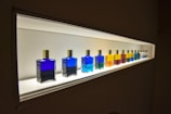 A row of colorful bottles with gold caps are displayed on a well-lit shelf. The bottles contain various vibrant liquids, ranging from deep blue to bright yellow, red, and green. The light from the shelf highlights the colors, creating a striking visual effect.