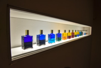 A row of colorful bottles with gold caps are displayed on a well-lit shelf. The bottles contain various vibrant liquids, ranging from deep blue to bright yellow, red, and green. The light from the shelf highlights the colors, creating a striking visual effect.