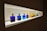 A row of colorful bottles with gold caps are displayed on a well-lit shelf. The bottles contain various vibrant liquids, ranging from deep blue to bright yellow, red, and green. The light from the shelf highlights the colors, creating a striking visual effect.